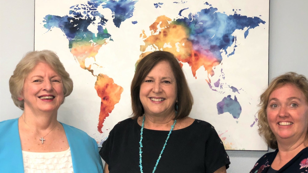Photo of Ann Vanvalen, Linda Adams, and Alma Thompson standing in front of a world map