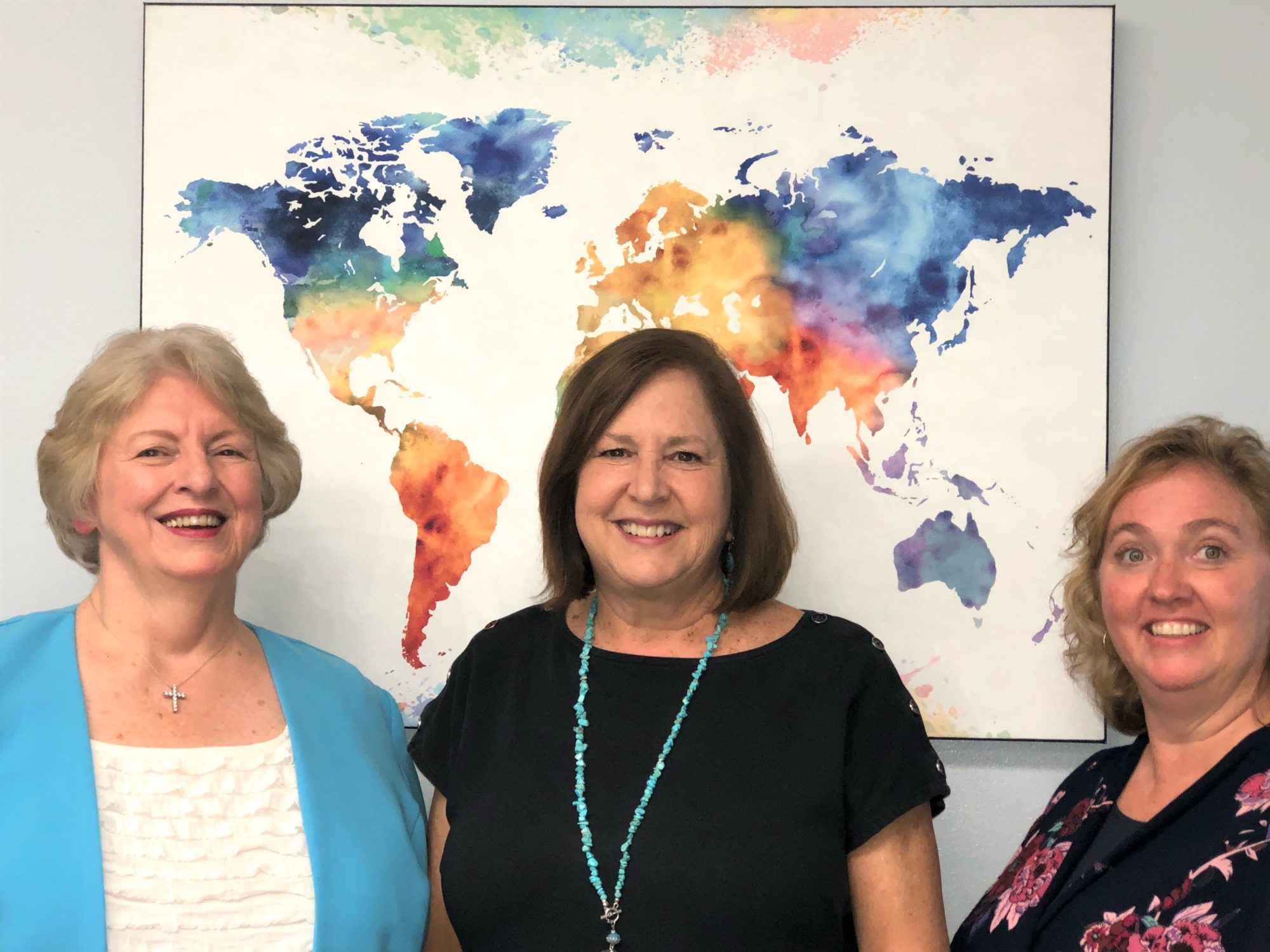 Photo of Ann Vanvalen, Linda Adams, and Alma Thompson standing in front of a world map